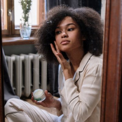 Young woman of color applying some skincare product to her face while sitting on the floor in front of a mirror.