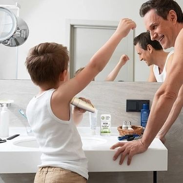 A small child with short hair stands in front of the bathroom sink applying MONA deodorant stick while their father looks on.