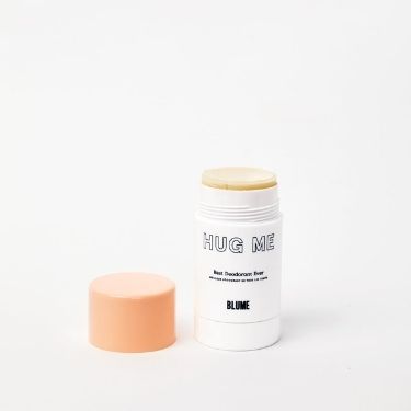 A stick of Blume's natural deodorant stick "Hug Me" with a simple white and black label. The orange top is off and you can see the yellowish colored stick.
