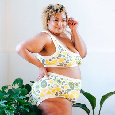 A Black plus size feminine presenting person standing sideways in front of plants with a hand on their hip and and another by their face smiles at the camera wearing organic cotton bralette and panty from Thunderpants